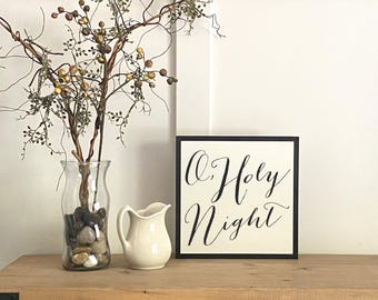 May include: A decorative arrangement featuring a clear glass vase filled with stones and branches with berries, a small white pitcher, and a framed sign with the text "O Holy Night" in elegant script. The items are on a wooden surface.