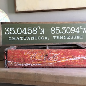 May include: A wooden sign with green paint and white lettering. The sign reads "35.0458°N 85.3094°W CHATTANOOGA, TENNESSEE".