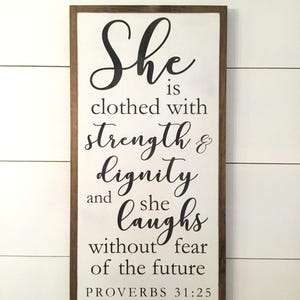 May include: A wooden sign with a white background and a brown frame. The sign features a black and white quote: "She is clothed with strength & dignity and she laughs without fear of the future. Proverbs 31:25".