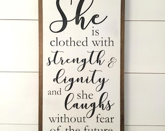 May include: A wooden sign with a white background and a brown frame. The sign features a black and white quote: "She is clothed with strength & dignity and she laughs without fear of the future. Proverbs 31:25".