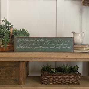 May include: A wooden sign with a green background and white text. The sign reads "But the fruit of the Spirit is love, joy, peace, patience, kindness, goodness, faithfulness, gentleness, and self control. Galatians 5:22-23".