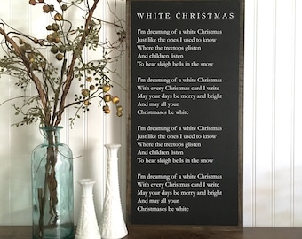 May include: A framed chalkboard sign with the words "WHITE CHRISTMAS" and the lyrics to the song. A clear glass vase with branches and berries, and two white decorative vases are on a wooden surface.