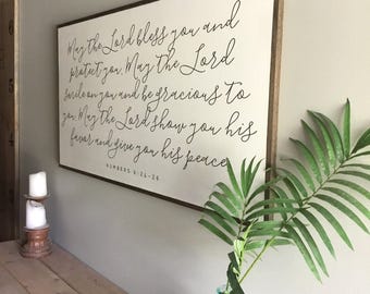 May include: A white wooden sign with a black cursive inscription that reads "May the Lord bless you and protect you. May the Lord smile on you and be gracious to you. May the Lord show you his favor and give you his peace. Numbers 6:24-26".
