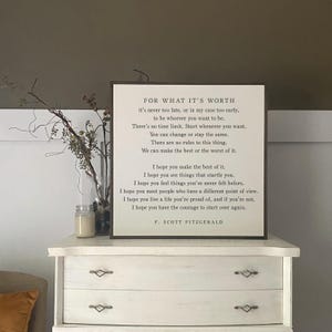 May include: A square wooden sign with a dark brown frame and a white background featuring the text of the F. Scott Fitzgerald poem For What It's Worth. The black printed text includes the full poem and the author's name at the bottom center.