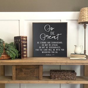 May include: Black framed sign with white text that reads "Go Be Great" and a bible verse from Joshua 1:9. The sign is on a wooden shelf with a book, a plant, and a basket.