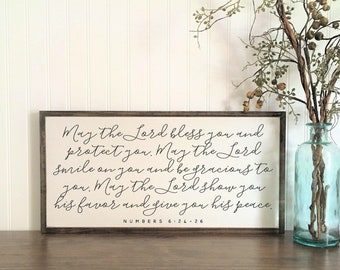 May include: A rectangular wooden sign with a dark brown frame and a white background. The sign features black cursive text that reads: May the Lord bless you and protect you. May the Lord smile on you and be gracious to you. May the Lord show you his favor and give you his peace. Numbers 6:24-26.