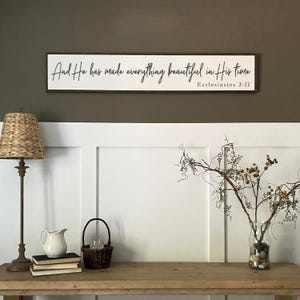 May include: A rectangular white wooden wall sign with a dark brown frame featuring the black cursive text And He has made everything beautiful in His time and the reference Ecclesiastes 3:11 printed in a simple black serif font.