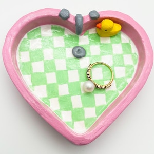 May include: A pink heart-shaped ceramic dish with a green and white checkered pattern inside. The dish has a small yellow rubber ducky and a gold ring with a pearl on top.