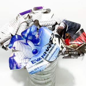 Puede incluir: Un ramo de páginas de periódico y revistas enrolladas en un jarrón de vidrio transparente. El ramo tiene forma de flor y tiene una página de revista azul y blanca con el texto "Evan Hansen" en ella.