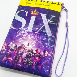 Puede incluir: Bolsa morada con cremallera con el musical "SIX", el título en letras blancas grandes y una corona dorada. La bolsa tiene una parte superior amarilla con las palabras "LAYBILL" y "BROOKS ATKINSON THEATRE". Se adjunta una correa de muñeca morada.