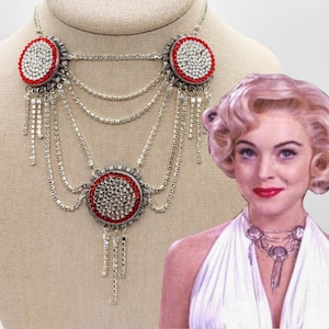 Op de afbeelding: Een meerrijige ketting met drie ronde hangers in de stijl van een kroonkurk, versierd met heldere en rode strass-steentjes. Het ontwerp bevat meerdere gedrapeerde zilverkleurige kettingen met heldere kristallen en bungelende kristallen franjes.