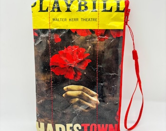 May include: A rectangular pouch with a red zipper and wrist strap. The pouch features a yellow top with the word "PLAYBILL" and "WALTER KERR THEATRE" printed on it. The main body of the pouch displays a red flower and the word "HADESTOWN".