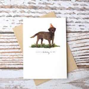 May include: A birthday card featuring a watercolor illustration of a brown Labrador Retriever wearing a red and white party hat. The text "Happy Birthday to You" is written below the dog.