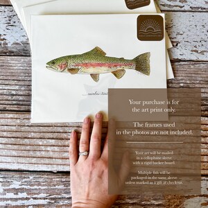 May include: Watercolor art print of a rainbow trout fish. The fish is depicted in shades of green, pink, and gold, with fine details. The print is on white paper, with the artist's signature and logo visible. The art print is packaged in a cellophane sleeve.
