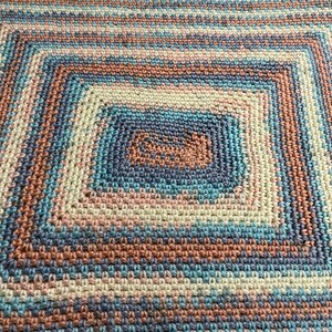 May include: A handmade crochet blanket with a square design. The blanket features concentric squares in shades of blue, peach, and cream. The texture of the crochet stitches is visible, creating a cozy and inviting appearance.