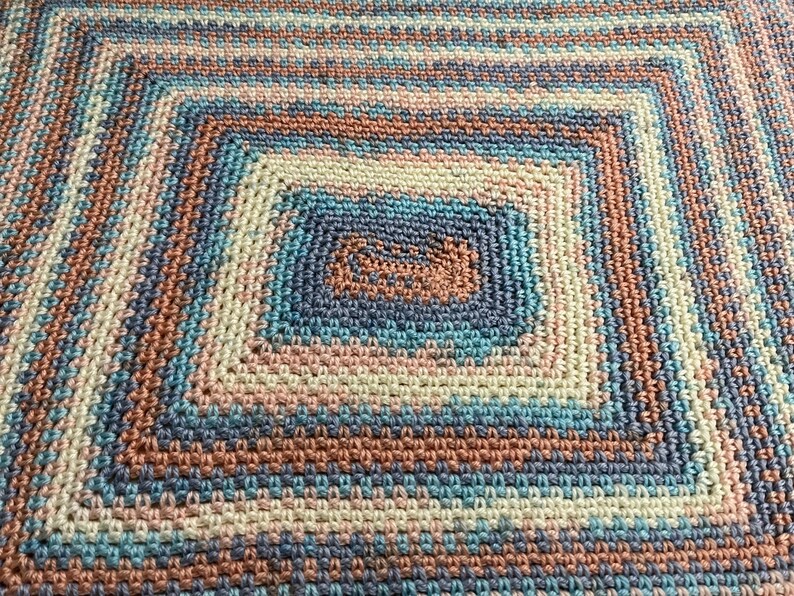 May include: A handmade crochet blanket with a square design. The blanket features concentric squares in shades of blue, peach, and cream. The texture of the crochet stitches is visible, creating a cozy and inviting appearance.