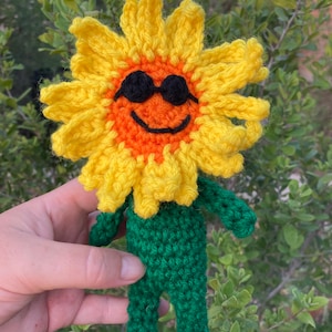May include: A crocheted sunflower with a smiling face and sunglasses. The sunflower is wearing a green crocheted outfit.