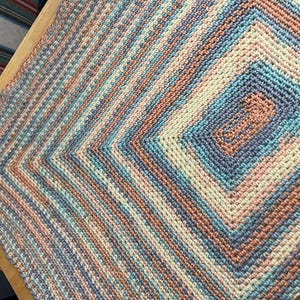 May include: A handmade crochet blanket in a square pattern. The blanket features a color scheme of light blue, peach, cream, and lavender. The design has a central square with concentric rectangles, surrounded by diagonal stripes.