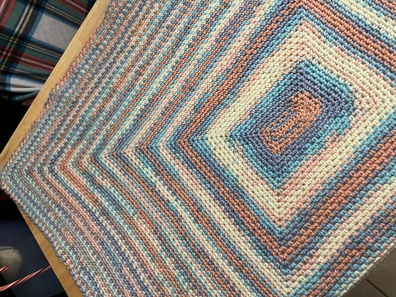 May include: A handmade crochet blanket in a square pattern. The blanket features a color scheme of light blue, peach, cream, and lavender. The design has a central square with concentric rectangles, surrounded by diagonal stripes.