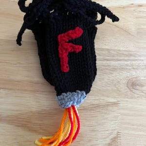 Op de afbeelding: Een zwarte gebreide tas met trekkoord en een rode "F" op de voorkant. De onderkant heeft een grijze rand en oranje, gele en rode garenstrengen. De tas staat op een houten oppervlak.