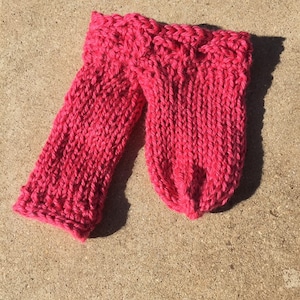 Könnte beinhalten: Ein Paar hellrosa gestrickte Fäustlinge. Die Fäustlinge sind aus dickem Garn mit einem strukturierten Strickmuster gefertigt. Die Bündchen haben ein dekoratives Zopfmuster. Die Fäustlinge liegen auf einer hellbraunen Oberfläche.