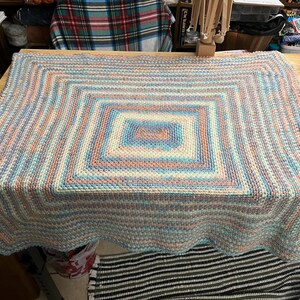 May include: A handmade, square-shaped blanket with a scalloped edge. The blanket features a geometric pattern with concentric squares in shades of blue, peach, and cream. The blanket is made of yarn and is displayed on a wooden table.