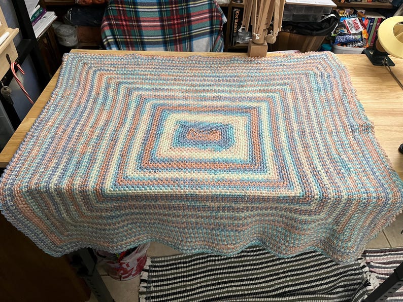May include: A handmade, square-shaped blanket with a scalloped edge. The blanket features a geometric pattern with concentric squares in shades of blue, peach, and cream. The blanket is made of yarn and is displayed on a wooden table.