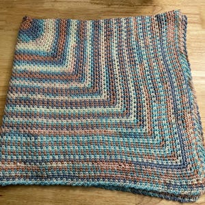 May include: A handmade, square crochet blanket in shades of blue, teal, peach, and cream. The blanket features a textured stitch pattern and a border in a darker teal. The blanket is folded on a wooden surface.