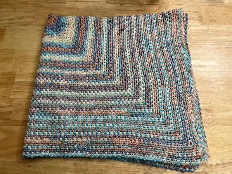 May include: A handmade, square crochet blanket in shades of blue, teal, peach, and cream. The blanket features a textured stitch pattern and a border in a darker teal. The blanket is folded on a wooden surface.