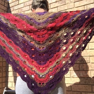 May include: A handmade, open-weave crochet shawl in a triangular shape. The shawl features a gradient of colors, including red, purple, brown, and tan. The design incorporates a scalloped edge and open, circular patterns throughout the fabric.