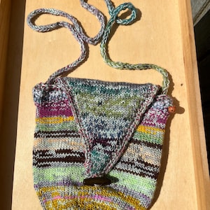 May include: A hand-knitted, colorful shoulder bag with a flap closure and a wooden button. The bag features a variety of colors, including green, purple, yellow, and blue. The strap is a braided cord. The bag is sitting on a wooden tray.