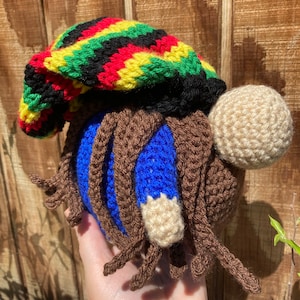 May include: A handmade crochet gnome with a colorful Rasta-style hat in red, yellow, green, and black. The gnome has brown dreadlocks, a blue body, and a beige beard. The gnome is a unique, whimsical decorative item.