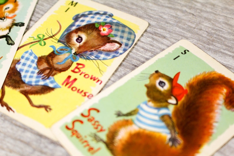 May include: Three illustrated cards from a children's game. The cards feature a brown mouse wearing a blue and white gingham dress and hat, a squirrel wearing a red and white striped shirt and a red hat, and a fox wearing a red and white striped shirt. The cards are labeled "Brown Mouse", "Sassy Squirrel", and "Foxy Fox".