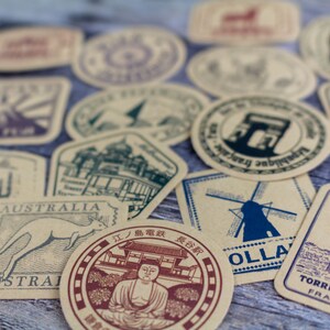 May include: A collection of vintage-style travel-themed stickers and labels. The designs feature iconic landmarks like the Eiffel Tower, the Leaning Tower of Pisa, and a windmill. The color palette includes shades of brown, blue, and red.