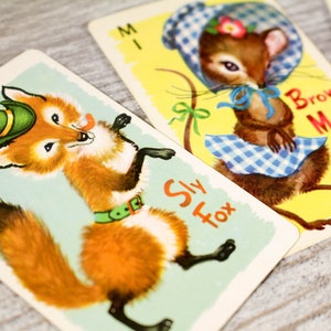May include: Two playing cards featuring illustrations of animals. The first card shows a sly fox wearing a green hat and a green belt. The second card shows a brown mouse wearing a blue and white gingham dress and a blue bow. The text on the first card reads "Sly Fox". The text on the second card reads "Brown Mouse".