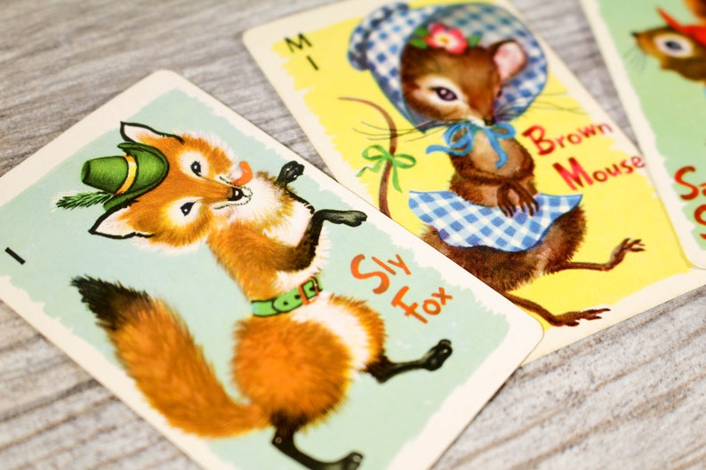 May include: Two playing cards featuring illustrations of animals. The first card shows a sly fox wearing a green hat and a green belt. The second card shows a brown mouse wearing a blue and white gingham dress and a blue bow. The text on the first card reads "Sly Fox". The text on the second card reads "Brown Mouse".