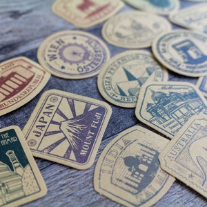 May include: A collection of vintage-style travel stickers with various designs and text. The stickers feature illustrations of landmarks and country names, including Japan, Australia, and Germany. The color palette includes shades of brown, blue, and purple.