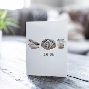 May include: A white card with a hand-drawn illustration of a loaf of bread, a bread proofing basket, and a jar of sourdough starter. The text "I LOAF YOU" is printed below the illustration.