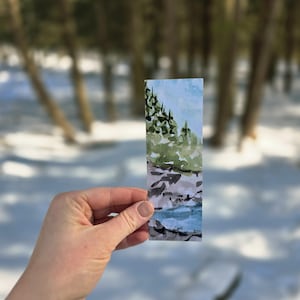 Könnte beinhalten: Ein rechteckiger Aquarell-Lesezeichen mit einer Landschaftsszene. Der obere Teil zeigt einen hellblauen Himmel und grüne Bäume. Die untere Hälfte zeigt einen Fluss mit grauen und schwarzen Felsen. Das Lesezeichen wird in einer Hand gehalten.