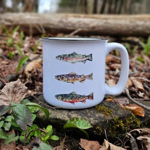 May include: White ceramic mug with a black rim and handle. The mug features three different illustrations of trout fish in various colors and sizes.