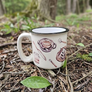 May include: Cream-colored enamel mug with a black rim and handle, decorated with watercolor illustrations of food items. The design includes a casserole dish, bread, and utensils. The mug is set outdoors on a bed of leaves and twigs.