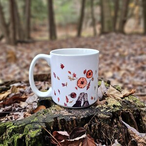 Könnte beinhalten: Weiße Keramik-Tasse mit einem Design aus Kürbissen, Blättern und einer Kaffeetasse. Die Tasse hat einen großen Henkel und ist vor einem verschwommenen Waldhintergrund platziert. Die Tasse steht auf einem Baumstumpf.