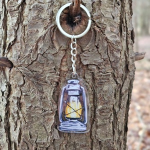 May include: A silver keychain with a clear acrylic charm featuring a hand-painted illustration of an oil lamp with a flame inside. The lamp is painted in shades of gray, blue, and yellow.