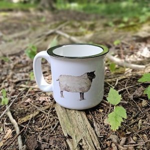 May include: White enamel mug with a black rim and handle. The mug features a watercolour illustration of a sheep. The mug is set on a bed of leaves and twigs.