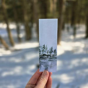 Könnte beinhalten: Ein rechteckiger Aquarell-Lesezeichen mit einer Winterlandschaft. Das Kunstwerk zeigt eine verschneite Szene mit immergrünen Bäumen, einem Gewässer und einem nebligen Himmel. Das Lesezeichen wird vor einem verschwommenen Waldbild gehalten.