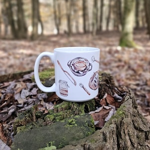 Peut inclure: Mug en céramique blanche avec des illustrations d'ustensiles de cuisine et d'aliments. Le motif comprend une tarte, un pot, une cuillère en bois et une miche de pain. Le mug est posé sur une souche d'arbre dans un cadre forestier.
