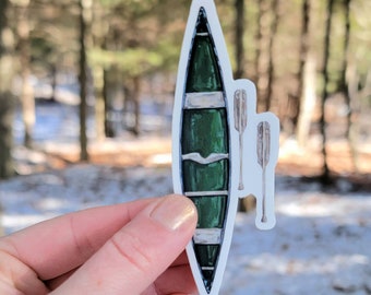 May include: A green canoe sticker with two white wooden paddles. The canoe has a white stripe down the middle and a white stripe around the top.