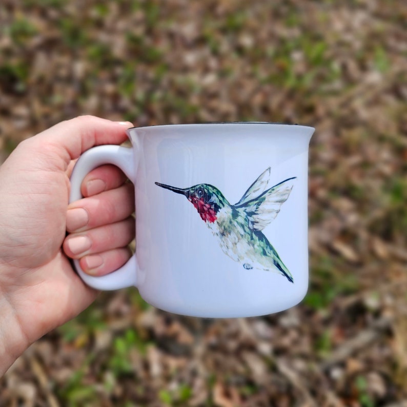 May include: White ceramic mug with a black rim and handle. A watercolor illustration of a hummingbird with green, red, and white feathers is printed on the mug.