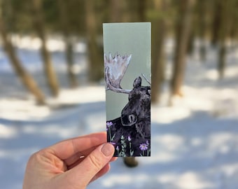 Puede incluir: Marcapáginas rectangular con una pintura de acuarela de un alce negro y gris con grandes astas. El fondo es verde pálido con flores moradas. El marcapáginas está sostenido en una mano.