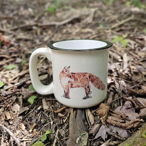 May include: Cream-colored ceramic mug with a black rim and handle. The mug features a watercolor illustration of a fox in shades of brown and orange. The mug is speckled and sits on a bed of leaves and twigs.